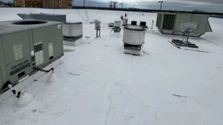 Commercial rooftop with multiple HVAC units, ventilation fans, and a satellite dish, all set on a white flat roofing membrane under a cloudy sky.