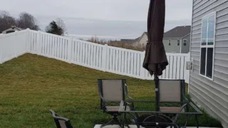 Backyard with a sloped lawn, white vinyl fence, patio table, and closed umbrella next to a house.