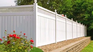Tall white vinyl privacy fence with decorative lattice top panels, set on a stone retaining wall next to green trees and red flowers