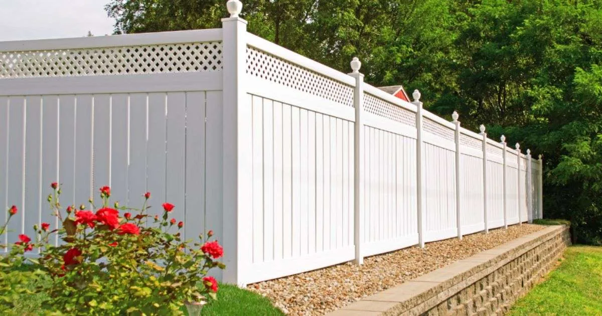 Tall white vinyl privacy fence with decorative lattice top panels, set on a stone retaining wall next to green trees and red flowers