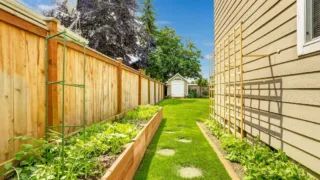 Narrow backyard with wooden fence, raised garden beds on one side, trellis on house wall, and a small shed in the distance.