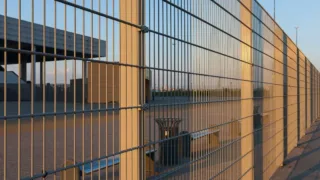 Close-up view of a tall metal security fence with vertical and horizontal bars, casting shadows at sunset.