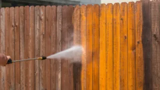 Person pressure washing a wooden fence, revealing clean, bright wood underneath the dirt.