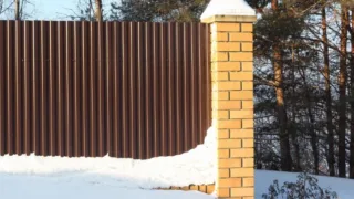 Brown corrugated metal fence with yellow brick pillar in a snowy forested area.
