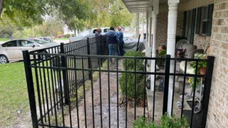 Workers installing a black metal fence in front of a brick house with a covered porch and outdoor furniture.