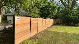 Long horizontal wooden fence with black metal posts running along a grassy backyard near trees.