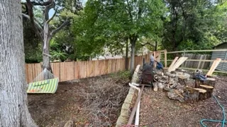 Backyard with new wooden fence, trees, a hammock, and stacked firewood near a small fire pit.