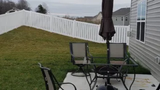 Backyard patio with table, chairs, and closed umbrella next to a house; white fence appears slanted due to uneven ground.
