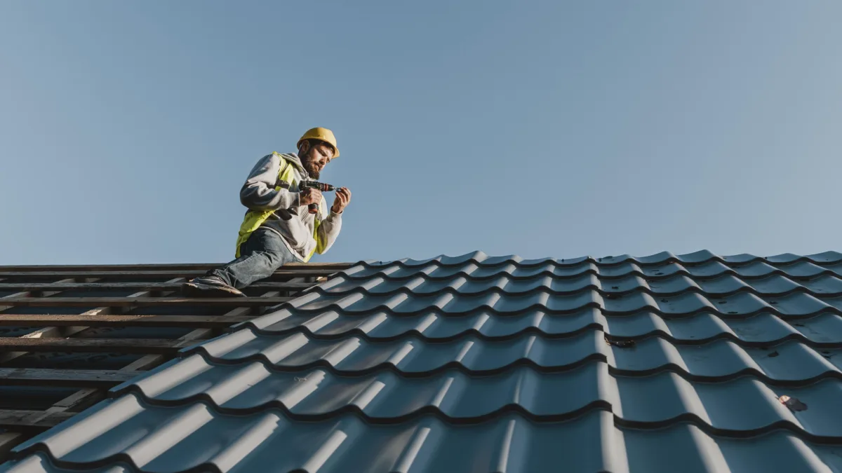 Professional roofer wearing safety gear installing metal roofing panels using a power drill on a residential roof under a clear blue sky.
