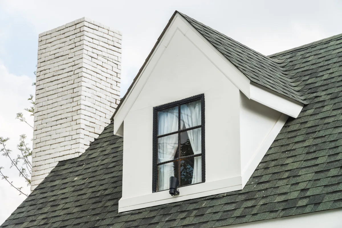Close-up view of a residential home with green asphalt shingle roofing, a white dormer window, and a white brick chimney.