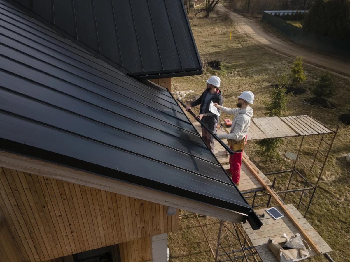 Two professional roofers wearing safety helmets installing black metal roofing panels on a wooden house using scaffolding during a construction project